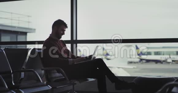 心情愉快的人坐在机场的椅子上等着他的航班然后他用笔记本电脑打字视频的预览图
