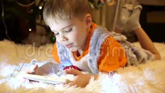 孩子躺在孩子们房间里柔软白色毯子上他正在智能手机上看动画片圣诞节视频的预览图