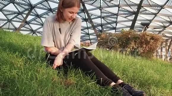 一个穿着灰色衬衫的少女坐在公园的草地上在阳光明媚的日子里读着一本带有讲稿的笔记本视频的预览图