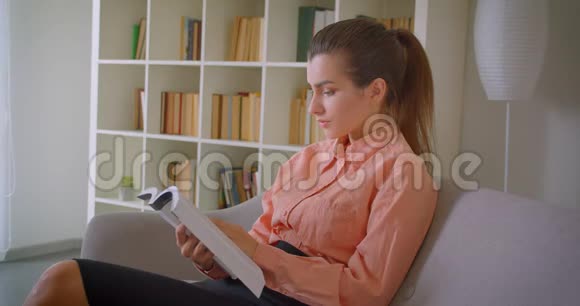 一位年轻漂亮的女商人坐在室内的沙发上看书的特写照片视频的预览图