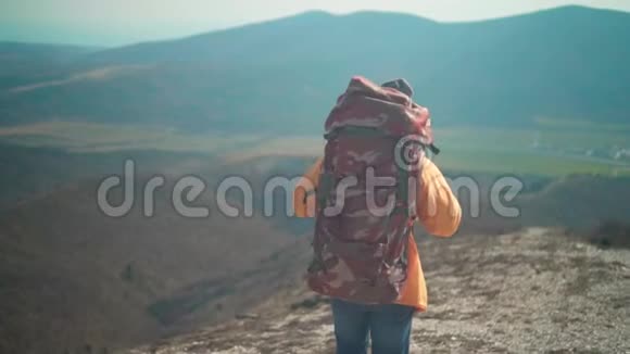 一个穿着黄色夹克蓝色牛仔裤和带着一个大型旅游背包的帽子的人穿过山脉向远处望去视频的预览图