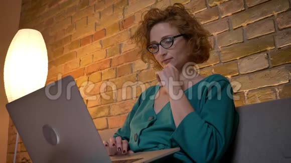 戴着眼镜的姜黄色卷发的白种人女孩体贴地用笔记本电脑工作双臂交叉在舒适的家里视频的预览图