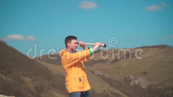一个穿着黄色夹克蓝色牛仔裤的年轻人坐在岩石上看着一个望远镜背景是山和天空视频的预览图