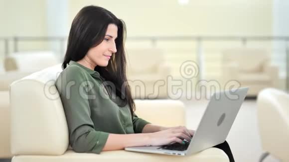 侧视微笑美丽的休闲女商人用笔记本电脑打字客厅视频的预览图