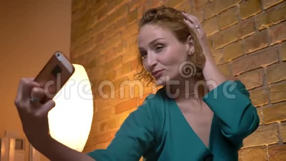 姜黄色卷发的白种人女孩用智能手机在舒适的家里拍自拍照片触摸头发视频的预览图