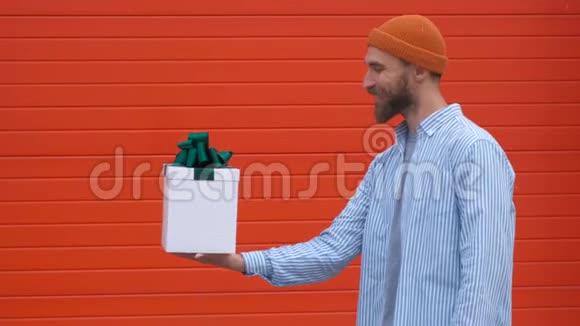 一位留着胡子和胡须的年轻时髦男士惊讶地拿着白色盒子红色背景上有礼物视频的预览图