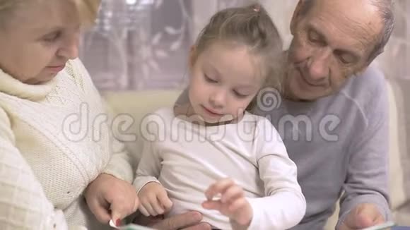 祖父和祖母正在读一本书他们的孙女坐在家里的沙发上家庭价值观视频的预览图