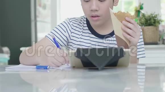 一个亚洲男孩在家做作业表情愉快手里拿着食物视频的预览图