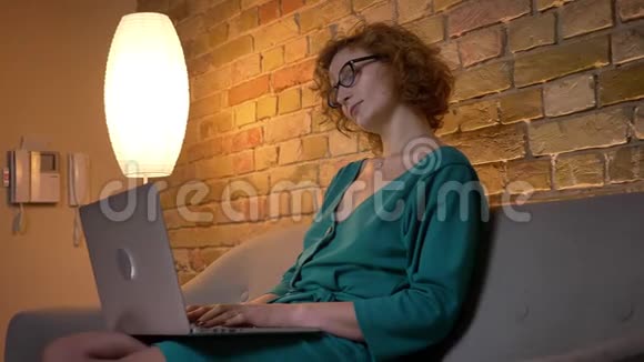在舒适的家中戴着笔记本电脑微笑着工作的戴着眼镜的姜黄色卷发白种人女孩的特写肖像视频的预览图