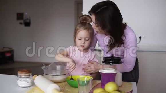 小女孩穿着粉红色的衬衫在厨房里和她的母亲一起在碗里搅拌面粉可爱的家庭爱的母亲吻她视频的预览图