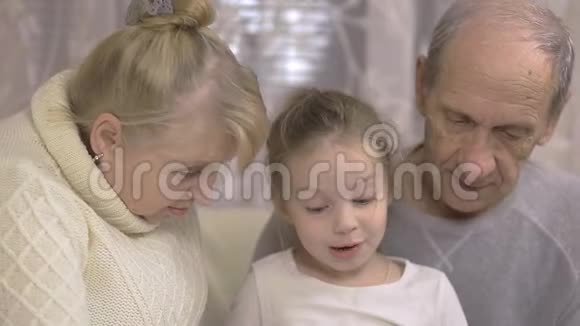 祖父和祖母正在读一本书他们的孙女坐在家里的沙发上家庭价值观视频的预览图