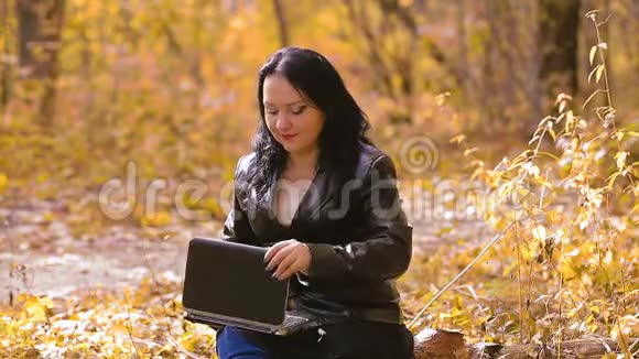 一个年轻的黑发女人在阳光明媚的秋日坐在公园里手提电脑远程工作视频的预览图