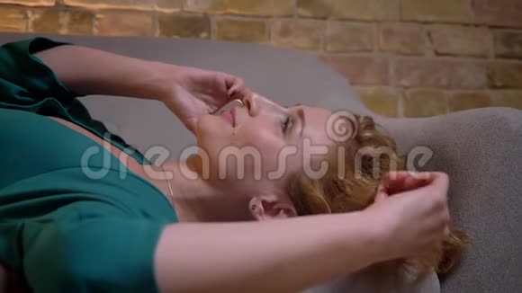 一幅姜黄色卷发的白种人女孩的特写肖像她躺在沙发上在智能手机上聊天把头发扭在一边视频的预览图