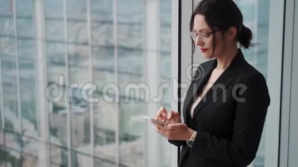 摩天大楼背景下一个商人在大窗户旁的肖像年轻女子使用手机视频的预览图