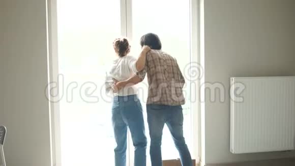 新婚夫妇幸福地拥抱在一间新公寓的窗户旁财产租赁住房购买青年贷款视频的预览图