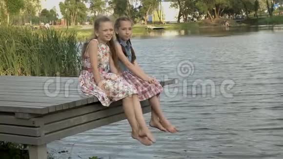 一个夏天的晚上两个快乐的小女孩坐在城市公园的河岸上坐在一座木桥上视频的预览图