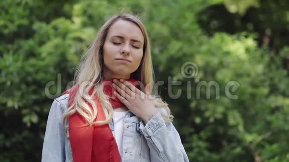 生病的年轻女子戴着红领巾感觉不好喉咙痛站在公园里痛苦视频的预览图
