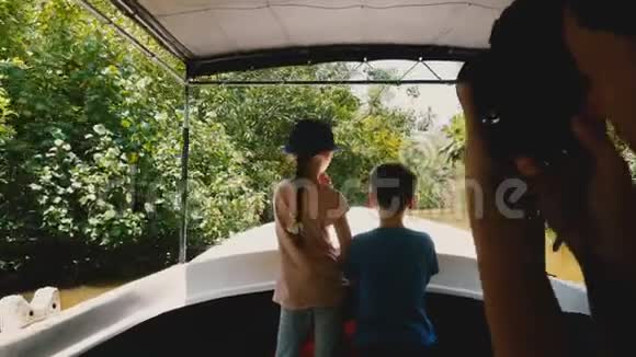 小快乐的女孩和男孩一起在狩猎之旅船上沿着异国情调的丛林河航行年轻的女人为他们拍照视频的预览图