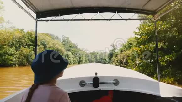 小快乐的女孩正坐在旅行船上沿着美丽的阳光明媚的丛林河缓慢航行水面上有茂盛的绿色植物视频的预览图