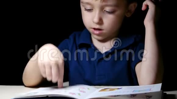 小孩用手指指着家里书中的图画晚上他坐在一张灯点亮的桌子旁视频的预览图