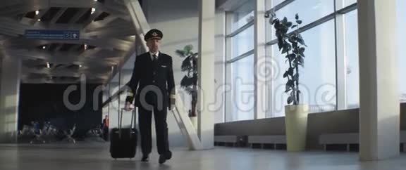 带手提箱戴帽子穿西装的成年飞行员在机场候机楼行走视频的预览图