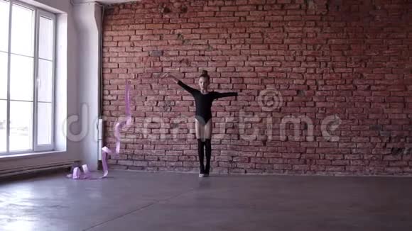 穿着黑色衣服的白人小女孩在带窗户和砖墙的工作室里用粉红色丝带表演体操视频的预览图