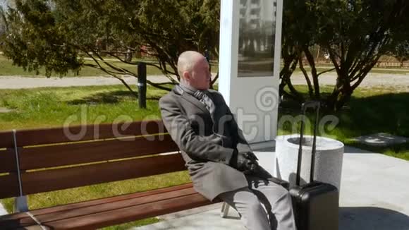 一个穿着灰色外套带着行李的优雅年轻人坐在长凳上休息着环顾四周旅行和商务视频的预览图