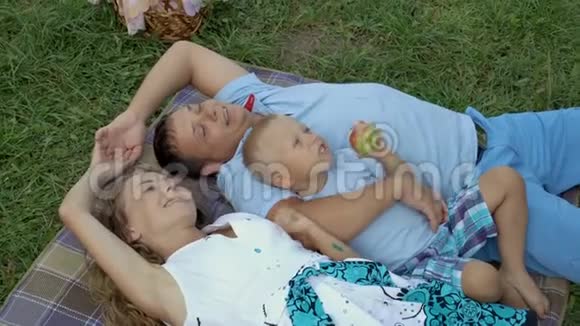 家庭在大自然中休息妈妈爸爸和小儿子躺在草地上的毯子上拥抱着看着天空男孩视频的预览图