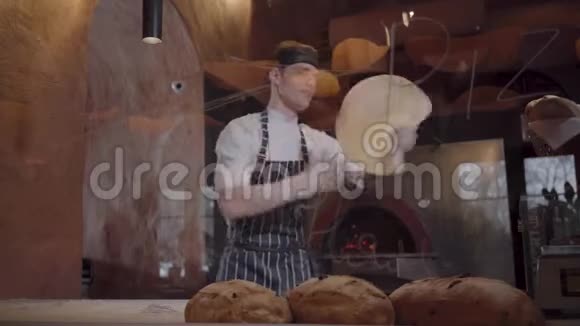 穿着厨师制服的年轻人在餐厅厨房里把披萨面团扔到玻璃后面技能视频的预览图