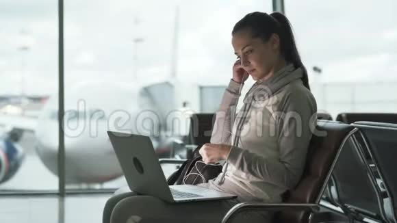 侧视女旅客戴上耳机在机场出发厅用笔记本电脑听音乐视频的预览图