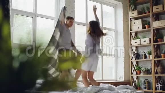 可爱的一对夫妇在床上跳舞毯子跳着一起笑着在晴朗的光线下度过周末的早晨视频的预览图