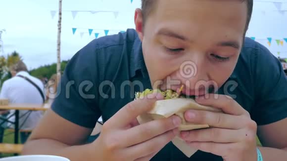 一个年轻人在一个节日或乡村集市上在露天咖啡馆里吃一个带有绿色面包的汉堡包或芝士汉堡视频的预览图