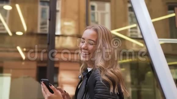 开朗的白种人女孩戴着时髦的眼镜在阳光明媚的日子里走在街上用智能手机发短信户外活动视频的预览图