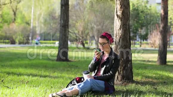 美丽年轻时尚的女人坐在公园的一棵树下喝着一杯咖啡和一部电话女孩自拍视频的预览图