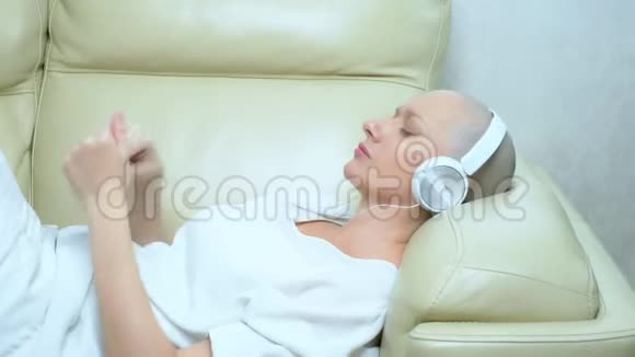 戴着耳机的漂亮秃顶女孩在家沙发上享受音乐视频的预览图