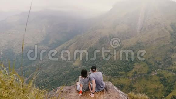 在斯里兰卡史诗般的山景中仰视高角度快乐浪漫的新婚夫妇在雨中坐在一起视频的预览图