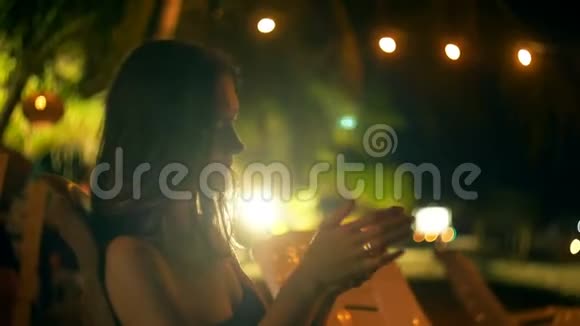 大气镜头耀斑拍摄的快乐年轻女商人坐在夜间休息室海滩酒吧椅子使用智能手机应用程序视频的预览图