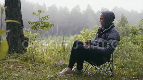 女徒步旅行者穿着夹克在森林里喝茶坐在自然露营的椅子上视频的预览图