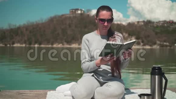 一个年轻的女人正在阳光下阅读一本书在春天的一天坐在一个湖的木墩上在大自然中放松视频的预览图