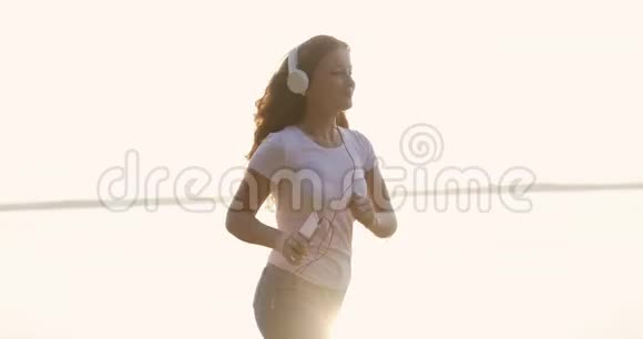 戴着白色耳机的红发女孩正在河边跑步用手机听音乐视频的预览图
