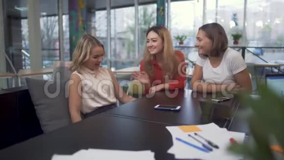三个笑着开玩笑的女孩坐在会议室的桌子上休息商务会议视频的预览图