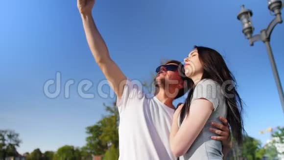 中镜头的快乐年轻夫妇自拍与智能手机同时站在公园与蓝天中等视频的预览图
