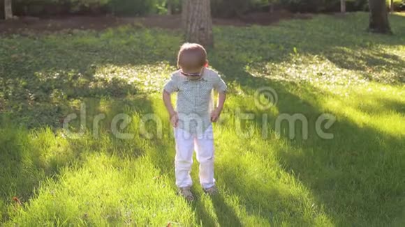 一个穿着白色裤子衬衫和太阳镜的小男孩站在绿草上视频的预览图