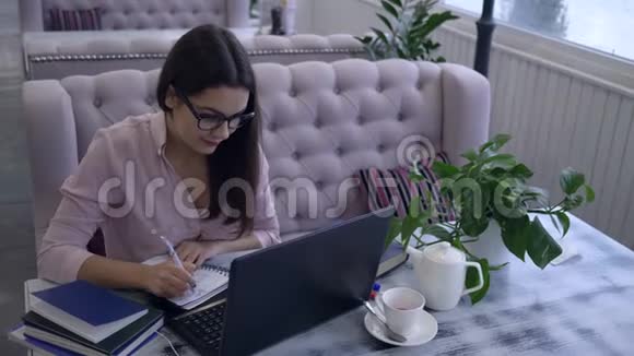 在咖啡馆的电子教育中戴眼镜的学生女孩用笔记本电脑写笔记用钢笔在笔记本上坐在桌旁视频的预览图