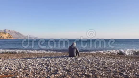 年轻人坐在靠近蓝色大海的海滩上扔着鹅卵石视频的预览图