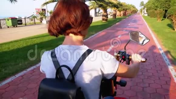 在度假小镇的红自行车道上女人骑着电动自行车骑着棕榈树视频的预览图