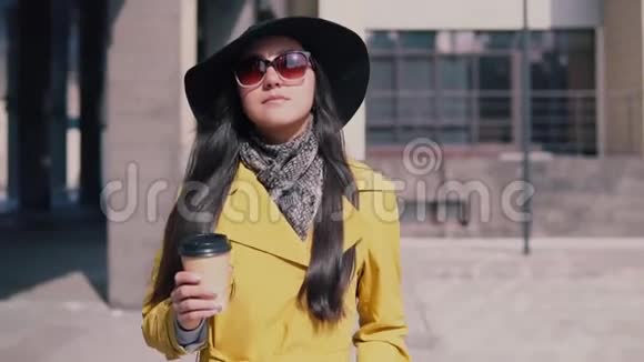 穿着黄色雨衣帽和眼镜的时髦女孩心情愉快地走在街上喝咖啡视频的预览图