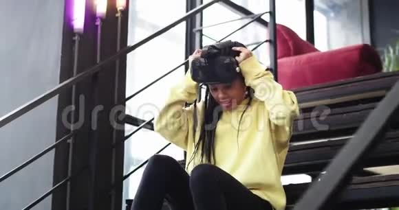 兴奋的非洲女士戴着虚拟现实眼镜坐在楼梯上在楼梯上玩得非常热情视频的预览图