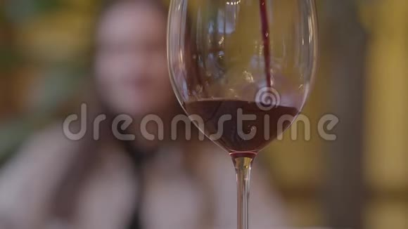 红酒慢慢地倒在前景的玻璃里关闭了年轻女子模糊的身影在背景中慢慢视频的预览图