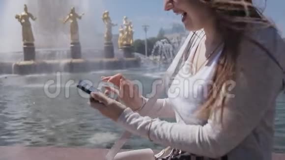 在炎热的夏天穿着米色西装坐在公园喷泉旁的智能手机的快乐女孩视频的预览图